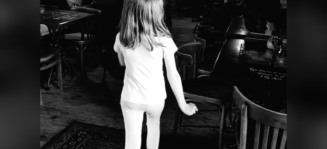 Kind zwischen leeren Tischen und Stühlen eines Cafés. Es schaut eine mechanische Spielorgel im Hintergrund an.